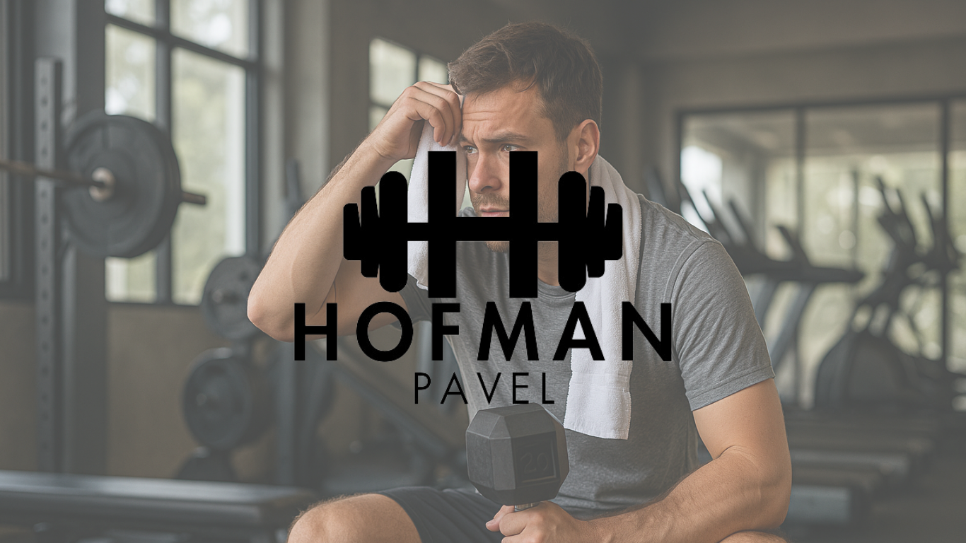Sportovec s ručníkem na krku drží činku a odpočívá po cvičení v moderním fitness centru – návrat k tréninku po pauze.